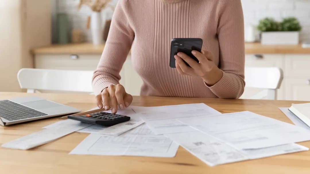 A woman sits at a table using a calculator and phone at home.