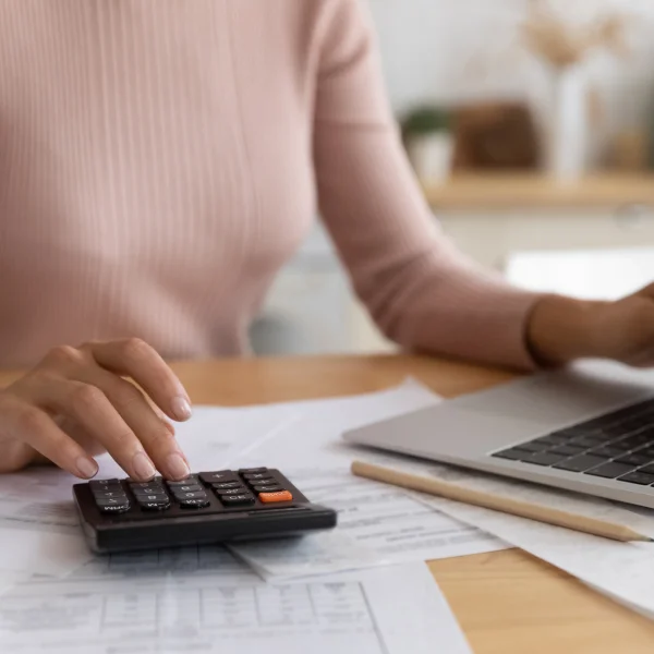 A woman calculates mortgage options using a calculator and laptop in her home office.
